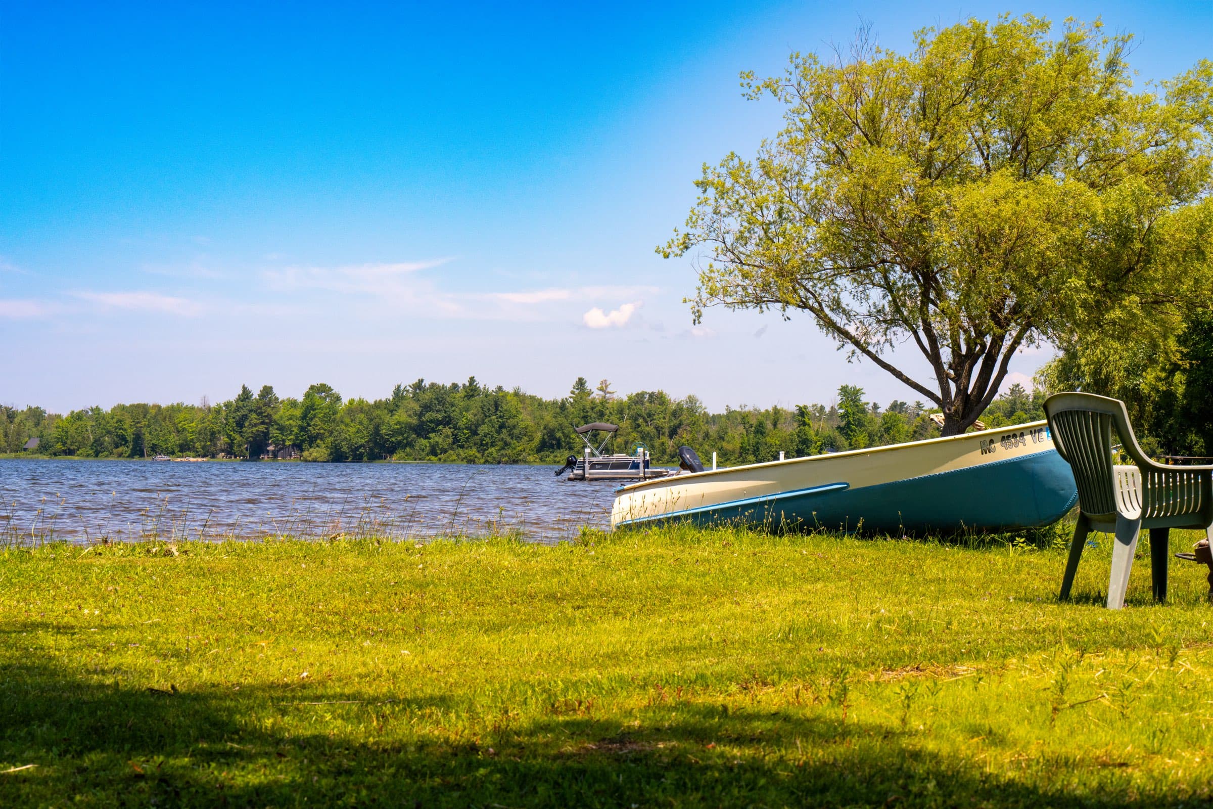 A canoe resting on the lawn beneath a willow, with a pontoon boat moored at the dock beyond