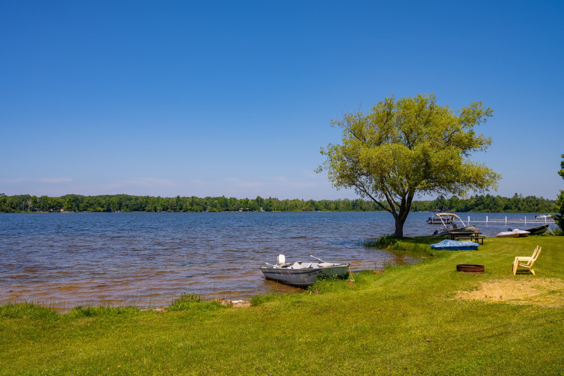 A rowboat pulled up on the grassy shore of Bass Lake beside a dock and an Adirondack chair