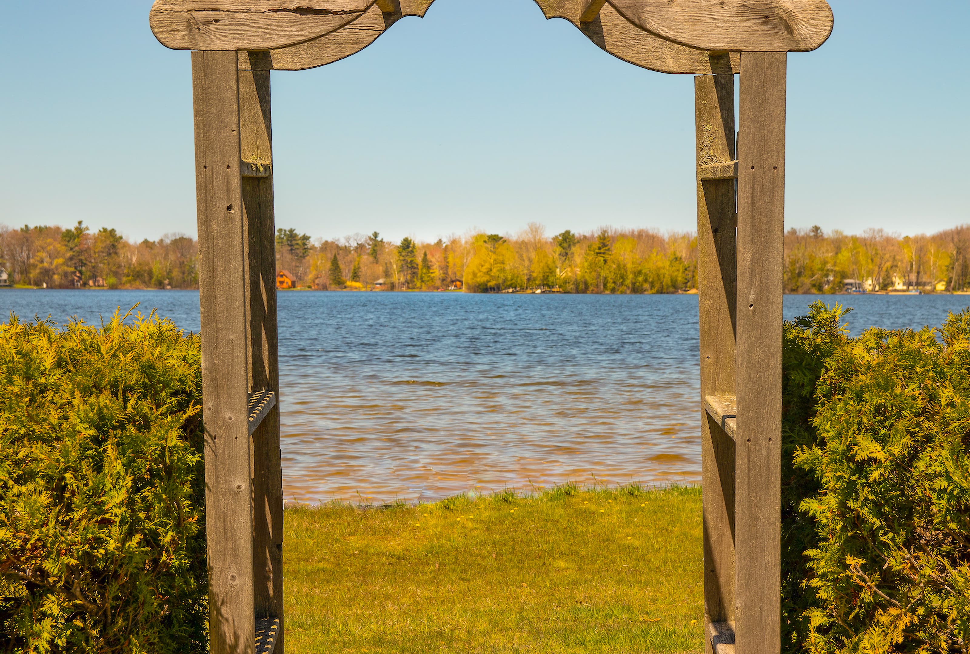 A wooden garden arbor framing the view across Bass Lake to the wooded far shore